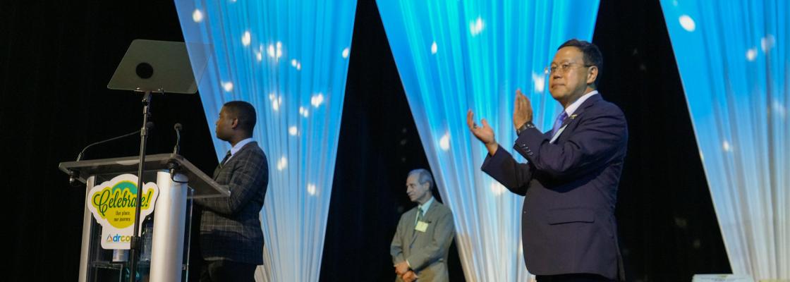 A person of Japanese descent wearing a suit stands on-stage and applauds an award winner.